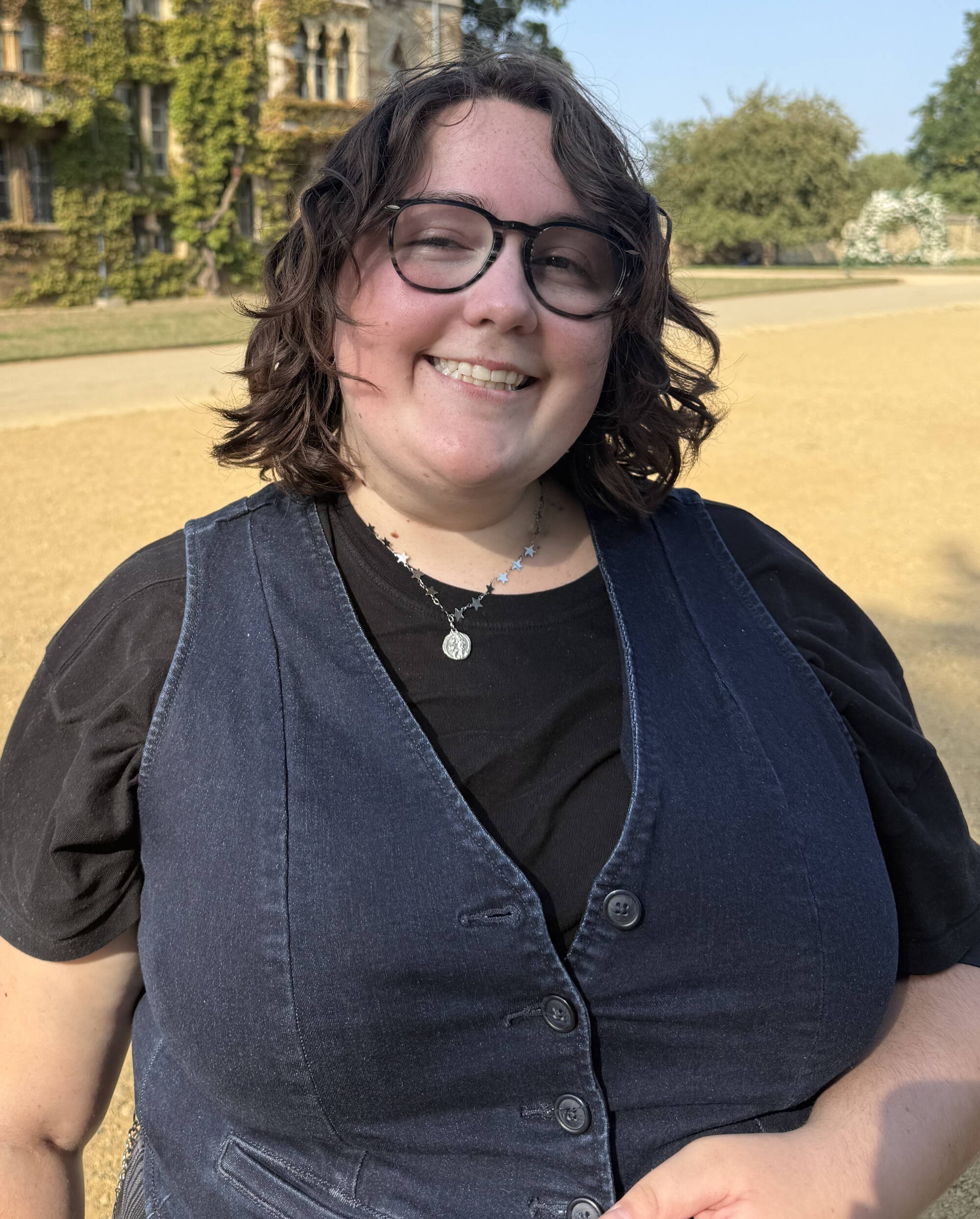 Woman with short, curly brown hair and glasses wearing a silver necklace and blue vest.